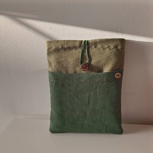 May include: A two-toned green fabric sleeve or case. The top section is a lighter olive green, and the bottom is a darker forest green. A wooden button and green cord secure the top flap. A small wooden button is on the front.