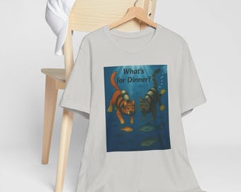 Op de afbeelding: Lichtgrijs t-shirt met een illustratie van twee katten die onder water duiken. De katten dragen duikmaskers en -flessen, met de tekst "What's for Dinner?" erboven. Vissen zwemmen in het blauwe water.