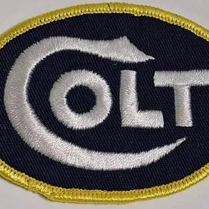 May include: Embroidered patch with the word "COLT" in white, stylized lettering. The patch is oval-shaped with a navy blue background and a gold border. The design features a distinctive, curved "C" and a simple "OLT".