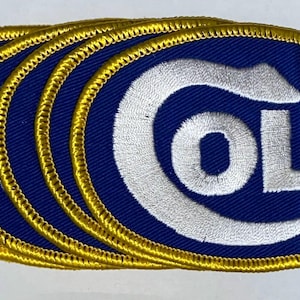 Puede incluir: Parches bordados con un diseño ovalado azul, con la palabra "COLT" en letras blancas y plateadas. Los parches tienen un borde dorado. Los parches están apilados uno encima del otro.