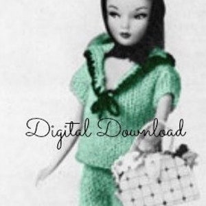 May include: A green knitted outfit for a doll, featuring a sailor-style top with a white collar and a matching skirt. The doll is holding a white and black polka dot bag.
