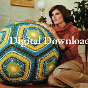 May include: A large, crocheted, multi-colored, geometric-shaped floor pillow. The pillow is made of blue, green, yellow, and white yarn. The pillow is sitting on a wooden floor. A woman is sitting on a couch behind the pillow.