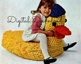 Puede incluir: Un pato amarillo de crochet con un pico rojo y un sombrero azul y blanco. Una niña está sentada en el pato, con un chaleco de crochet blanco y rosa y medias blancas.