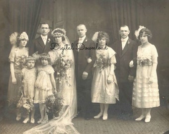 May include: A black and white vintage photograph of a wedding party. The bride is wearing a white dress with a long veil and a bouquet of flowers. The groom is wearing a suit and tie. The bridesmaids are wearing dresses and the groomsmen are wearing suits. The group is standing in front of a backdrop of curtains.