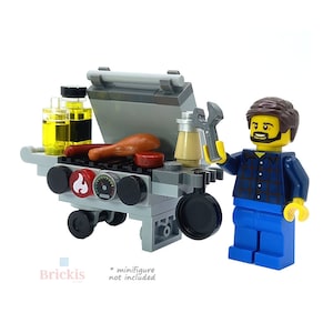 Op de afbeelding: Een grijze en zwarte speelgoedgrill met een rode vlam en een zwarte wijzerplaat. De grill heeft twee flessen olie, een hotdog en een spatel. Een geel Lego-poppetje met een blauw geruit overhemd en een blauwe broek staat naast de grill.