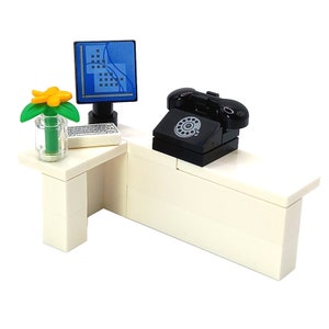 Könnte beinhalten: Ein weißer Schreibtisch mit einem schwarzen Drehwahltelefon, einem Computermonitor und einer Tastatur. Eine kleine Topfpflanze mit einer gelben Blüte steht auf dem Schreibtisch.