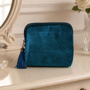 May include: A square-shaped teal velvet cosmetic bag with a shimmering texture. The pouch features a dark zipper closure and a matching teal fabric tassel attached to the zipper pull with a gold-toned metal cap, providing a simple and functional design.