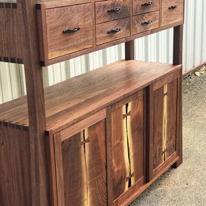 K&ouml;nnte beinhalten: Ein hoher Holzschrank mit einem Regal und sechs Schubladen. Der Schrank hat zwei T&uuml;ren mit einer Holzmaserung. Der Schrank ist aus Walnussholz.