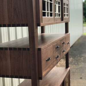 K&ouml;nnte beinhalten: Ein hoher Holzschrank mit dunkelbrauner Oberfl&auml;che. Der Schrank hat zwei Glast&uuml;ren mit einem Gittermuster und eine Reihe von Schubladen darunter. Der Schrank ist aus massivem Holz gefertigt und hat einen traditionellen Stil.
