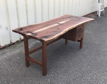 May include: A solid wood desk with a live edge top and three drawers. The desk is made of dark brown wood and has a natural, rustic look.