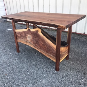 May include: A dark brown wooden table with a live edge top and a unique, curved shelf underneath. The table is made of solid wood and has a natural, rustic finish.