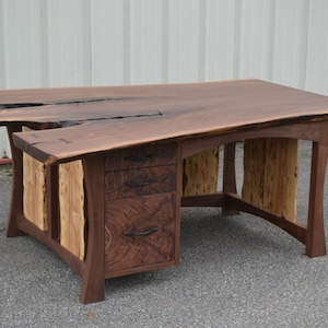 May include: A large, dark wood desk with a live edge top and three drawers. The desk has a unique design with a mix of wood types and a natural edge.