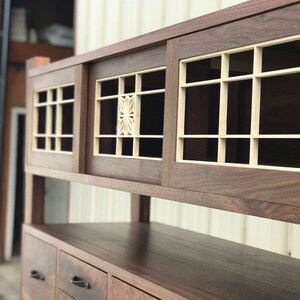 K&ouml;nnte beinhalten: Ein Holzschr&auml;nkchen mit zwei Schiebet&uuml;ren, die ein geometrisches Muster aufweisen. Der Schrank hat ein oberes Regal und drei Schubladen mit schwarzen Griffen.