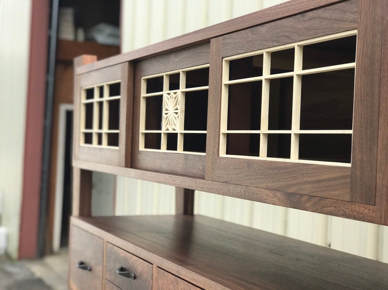 K&ouml;nnte beinhalten: Ein Holzschr&auml;nkchen mit zwei Schiebet&uuml;ren, die ein geometrisches Muster aufweisen. Der Schrank hat ein oberes Regal und drei Schubladen mit schwarzen Griffen.