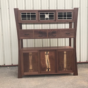 K&ouml;nnte beinhalten: Ein hoher, dunkler Holzschrank mit drei Regalen. Das obere Regal hat drei Glast&uuml;ren mit einem geometrischen Muster. Das mittlere Regal hat vier Schubladen mit schwarzen Griffen. Das untere Regal hat drei T&uuml;ren mit einer Holzmaserung.