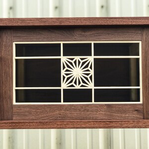 K&ouml;nnte beinhalten: Ein Holzschr&auml;nkchen mit einer Glast&uuml;r, die ein geometrisches Muster in der Mitte zeigt. Der Schrank ist aus dunklem Holz mit einer Einlage aus hellem Holz.