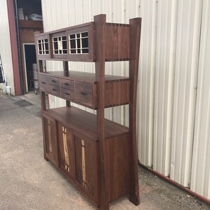K&ouml;nnte beinhalten: Ein hoher, dunkelbrauner Holzschrank mit Regalen, Schubladen und T&uuml;ren. Der Schrank hat ein einzigartiges Design mit einer gew&ouml;lbten Vorderseite und einem dekorativen Holzmaserungsmuster.