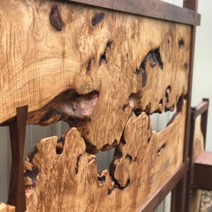 May include: A wooden headboard with a natural wood finish. The headboard features a unique design with a large, irregular opening in the center. The opening is surrounded by a frame of darker wood.