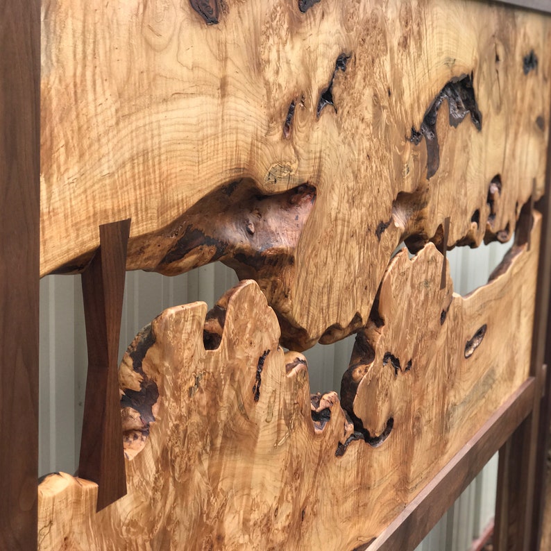May include: A wooden headboard with a natural wood finish. The headboard features a large, irregular shape cut out of the center, revealing the wall behind it. The headboard is framed with a dark wood border.