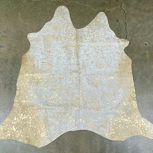 May include: White cowhide rug with gold accents. The rug has a natural, distressed look and is perfect for adding a touch of rustic elegance to any room.