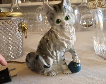 May include: A ceramic cat figurine with grey and white markings, green eyes, and a blue ball. The cat is seated and holding the ball. Glass bottles and jars are visible in the background.