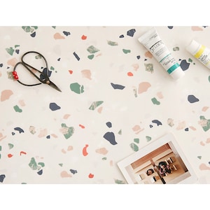 May include: A white background with a terrazzo pattern in shades of pink, green, and blue. There are two tubes of skincare products, a pair of scissors, and a photograph of two people.