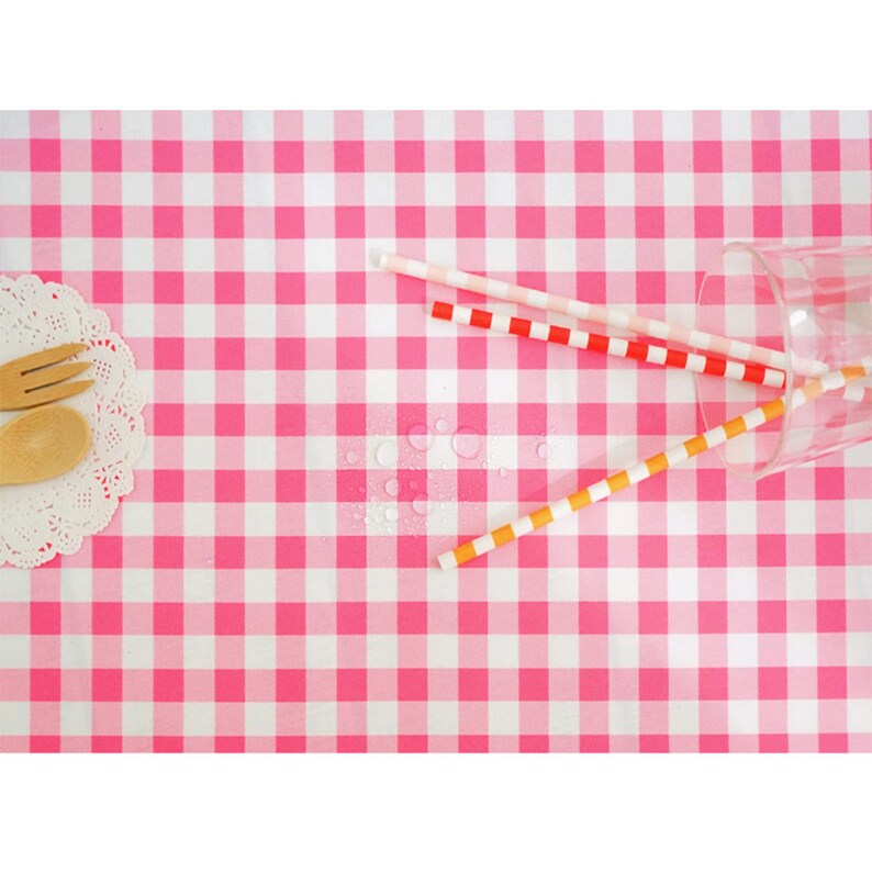 May include: Pink and white checkered tablecloth with three colorful paper straws and a glass on top. Water droplets are visible on the tablecloth.