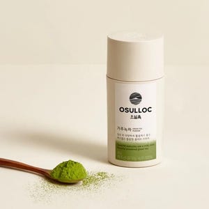 Könnte beinhalten: Ein weißer zylindrischer Behälter mit grünem Tee-Pulver mit dem Markennamen "OSULLOC" und dem Text "Green Tea Powder" in Koreanisch und Englisch. Der Behälter steht auf einer weißen Oberfläche mit einem Holzlöffel gefüllt mit grünem Tee-Pulver.