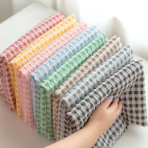 May include: A selection of folded cloths in a range of colours, including red, yellow, pink, blue, green, brown, and grey. Each cloth features a checkered pattern. The cloths are held by a person's hands, displaying the variety of colours.