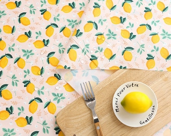 May include: A pink tablecloth with a lemon and leaf pattern. The tablecloth is folded over a wooden cutting board with a fork and a small white plate with a lemon and the text "Merci pour votre gentillesse".