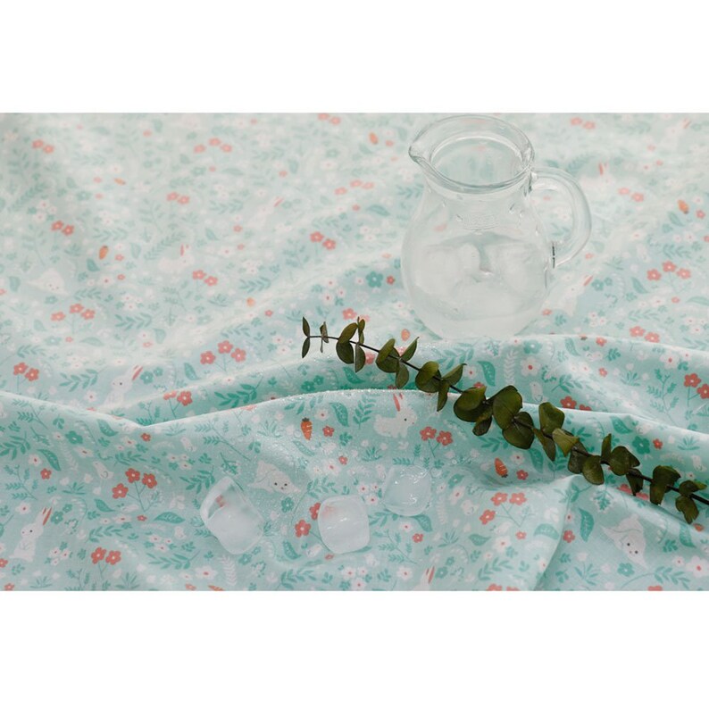 May include: A light blue fabric with a floral pattern featuring small white bunnies and pink flowers. A clear glass pitcher and a sprig of green eucalyptus leaves are on the fabric.