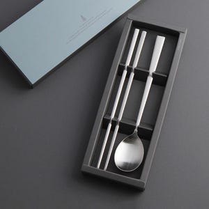 May include: A set of silver chopsticks and a spoon in a black box. The chopsticks are arranged in a row, and the spoon is placed on top of them. The box is made of cardboard and has a simple design.