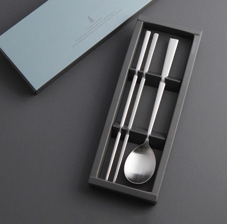 May include: A set of silver chopsticks and a spoon in a black box. The chopsticks are arranged in a row, and the spoon is placed on top of them. The box is made of cardboard and has a simple design.