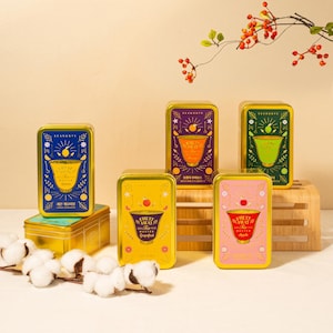 May include: A collection of colorful rectangular metal tins with gold trim. Each tin features a unique design with a fruit illustration and text. The tins are arranged on a wooden stand and a gold box, with cotton buds in the foreground.