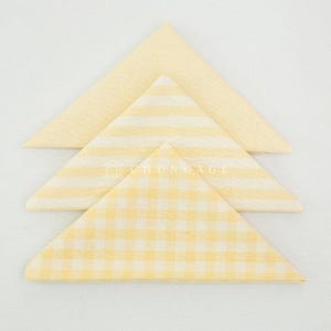 May include: Three folded cloth napkins in a triangle shape. The top napkin is a solid pale yellow, the middle one has yellow and white stripes, and the bottom one has a yellow and white gingham pattern. The napkins are arranged on a white surface.