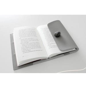 May include: Open book with white pages inside a gray leather book cover. The cover has a flap with a small, textured, gray stone closure. The book is on a white surface.