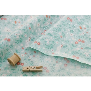 May include: A light blue fabric with a white bunny and pink flower pattern. The fabric is folded and has water droplets on it. There is a wooden spool of thread and a wooden clothespin on the fabric.