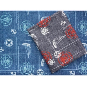 Op de afbeelding: Twee stofstalen met nautische patronen. De bovenste staal is een donkerblauwe denim met een wit kompas-, anker- en reddingsboei-ontwerp. De onderste staal is een donkergrijze stof met een rood en wit kompas-, anker- en bloemachtig ontwerp. De tekst "Cotton Club marinship" wordt op beide stalen herhaald.
