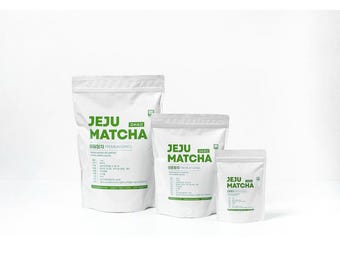 Puede incluir: Tres bolsas blancas de té Jeju Matcha, cada una con texto verde. La bolsa más grande está a la izquierda, seguida de una mediana y una pequeña. El texto incluye "JEJU MATCHA" y otro texto coreano. Las bolsas están selladas.