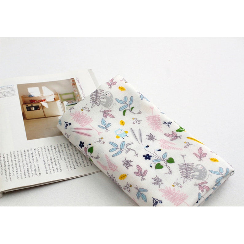 May include: White fabric with a repeating pattern of pink, green, blue, and yellow leaves, flowers, and small animals. The fabric is folded in half and lying on top of a magazine.