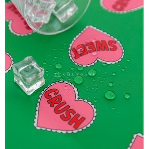 Op de afbeelding: Een groene ondergrond met roze hartvormige ontwerpen met de rode woorden "CRUSH" en "SWEET". Een glas met ijsblokjes staat linksboven. Waterdruppels zijn verspreid over het oppervlak.