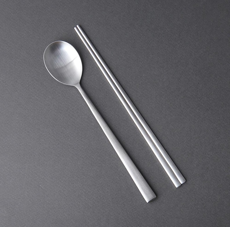 May include: A set of silver chopsticks and a spoon. The chopsticks are laid out parallel to each other, while the spoon is placed diagonally across them.
