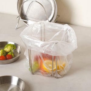 Può includere: Un cestino in filo di acciaio inossidabile contenente un sacchetto di plastica trasparente pieno di frutta colorata. Fette di limone, pomodori rossi e broccoli verdi sono in vassoi di acciaio inossidabile. Un portapiatti in acciaio inossidabile è sullo sfondo.