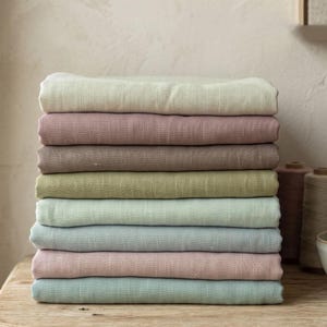 May include: A stack of eight folded fabric pieces in various muted pastel colours including cream, dusty rose, taupe, olive green, mint, light blue, soft pink, and seafoam green. Each piece of fabric displays a visible woven texture throughout.