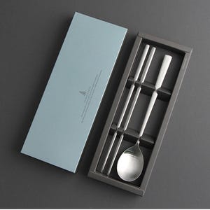 May include: A set of silver chopsticks and a spoon in a gray box with a light blue lid. The box has a small tree logo and text on the lid.
