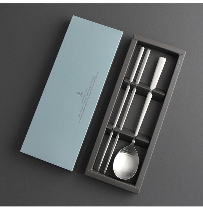 May include: A set of silver chopsticks and a spoon in a gray box with a light blue lid. The box has a small tree logo and text on the lid.