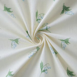 Può includere: Tessuto bianco sporco con un motivo ripetuto di fiori a forma di campana blu chiaro e foglie verdi. Il tessuto è piegato, creando un effetto a spirale. Il design floreale è delicato e sembra dipinto a mano.