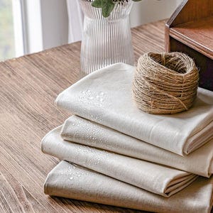 Op de afbeelding: Een stapel van vijf witte stoffen vierkanten met waterdruppels erop. De stof is netjes gevouwen en ligt op een houten tafel. Er is een bol garen en een glazen vaas met groene planten op de achtergrond.