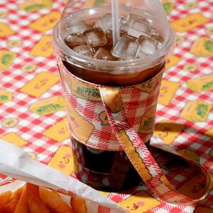 May include: A clear plastic cup filled with iced coffee and ice cubes, with a straw. The cup is wrapped in a patterned sleeve with a strap. A basket of french fries sits in the foreground, on a red and white checkered tablecloth.