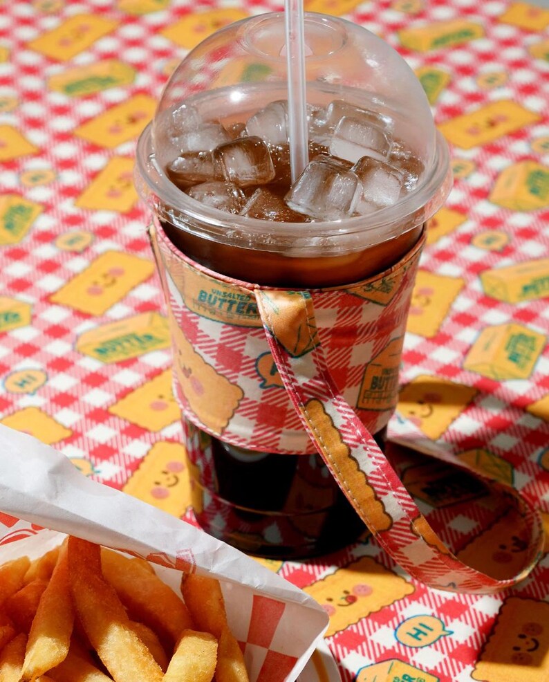 May include: A clear plastic cup filled with iced coffee and ice cubes, with a straw. The cup is wrapped in a patterned sleeve with a strap. A basket of french fries sits in the foreground, on a red and white checkered tablecloth.